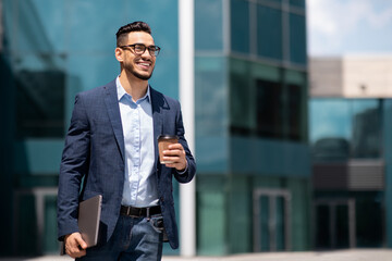 Cheerful middle eastern young businessman in glasses with laptop and coffee to go walking by street...