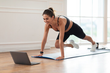 Morning Routine Concept. Cheerful slim lady in black sportswear and sneakers doing mountain climber exercise on yoga mat and watching online tutorial, looking at computer screen on floor at home gym