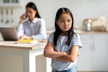 Working mom doing online job, her offended daughter feeling lonely and sad, standing in kitchen...