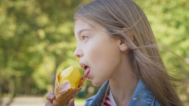 Side view of little Caucasian girl with long hair licking ice cream in waffle cone. Child in denim jacket enjoying cold dessert outdoors on sunny day in green summer park. Eating sweets.
