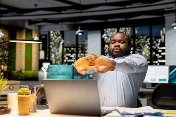 African american stiff man takes a break stretching muscles in the office, feeling overworked after hours and struggling to stay awake. Sleep deprivation and work tension due to workload.