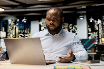 Black office employee checking a notification connected on socials while he does overtime at midnight, checking smartphone at work after hours. Young man reading text messages.