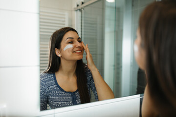 Beautiful young woman applying moisturizing face cream in front of a bathroom mirror