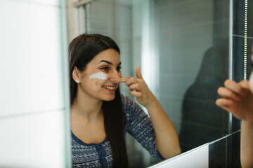Beautiful young woman applying moisturizing face cream in front of a bathroom mirror