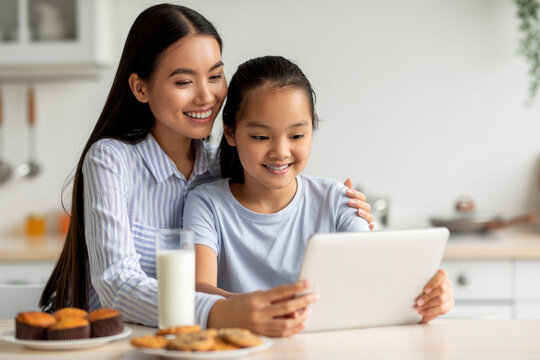 Happy asian mother and her cute daughter using digital tablet, looking for new recipes while sitting at kitchen, free space. Young mom and child spending time together at home - Powered by Adobe