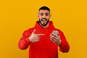 A man stands against a yellow background, smiling widely. He holds a smartphone in one hand and points at it with the other hand. His casual attire suggests a relaxed moment.