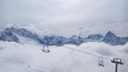 Chairlift above clouds with snow-covered mountain peaks at Dombay ski resort, Caucasus Mountains, Russia. High-altitude winter scene with alpine ridges and soft overcast sky