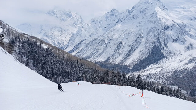 Skier descending a snowy slope at Dombay ski resort with dramatic Caucasus mountain peaks and dense pine forest. Winter sports scene in high-altitude alpine landscape