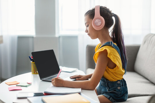 Online Education. Profile side view of focused schoolgirl with dyed colorful hair in headphones studying at home, writing in copybook or diary, attending webinar, e-learning during pandemic concept - Powered by Adobe