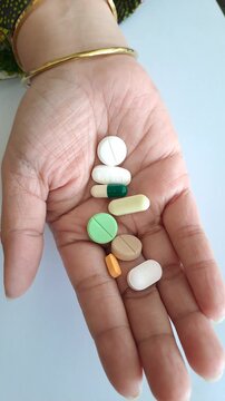 Close-up of an Asian woman's hand holding a variety of colorful pills and capsules. Concepts of medication, health treatment, supplement intake, pharmacy, and medical overdose risk awareness.