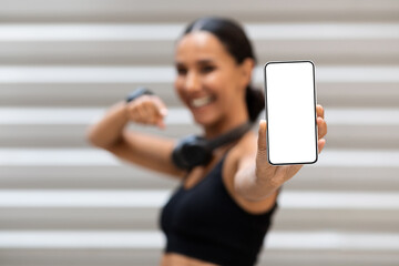 Satisfied pretty millennial arabic lady athlete with headphones pointing finger at phone with blank screen on wall background. App for training, great sale, website, offer and ad at covid-19 pandemic