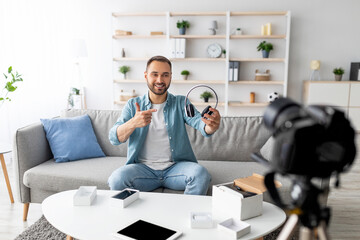 Cheery male vlogger recording video review of wireless headphones at home. Positive young blogger...