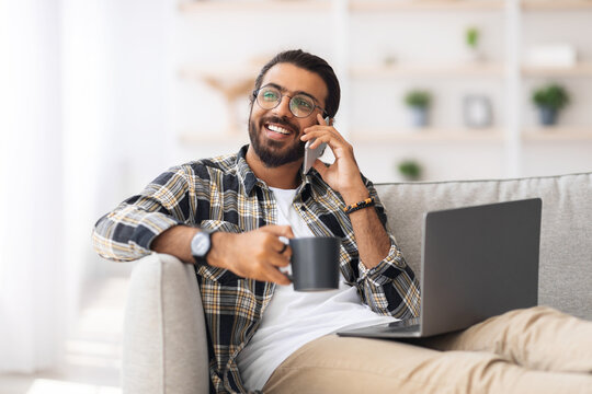 Relaxed arab guy sitting on sofa with laptop and mug with hot drink, having phone coversation while chilling at home, looking at copy space and smiling. Domestic entrertainment and technologies