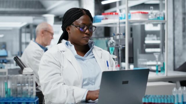 Portrait of cheerful scientist using laptop software in laboratory, driving treatment discovery. Jolly woman using bioinformatics application on notebook in sterile lab, camera B