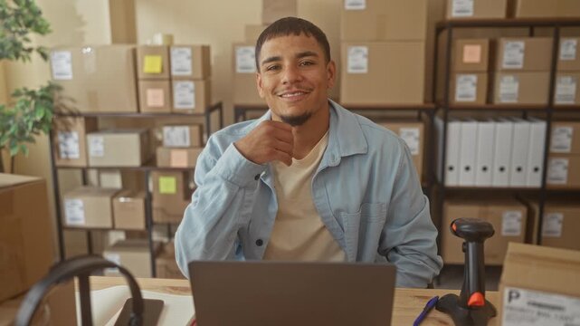 Man with hand on chin at desk with laptop and barcode scanner, surrounded by boxes and shelves in building; small business confidence.