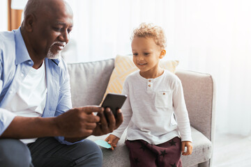 A grandparent and a young child sit on a couch in a living room. They are looking at a phone together, sharing a moment and enjoying each other's company.
