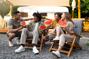 Joyful diverse friends chatting near camper van, eating yummy watermelon, sitting in lounge chairs,...