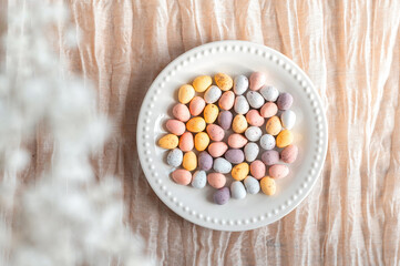 Colorful Easter chocolate eggs in a plate with flowers on a beige background, top view.