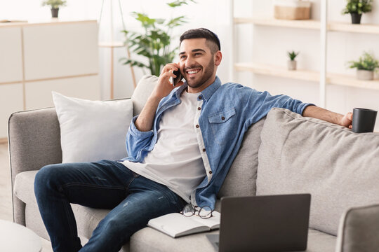 Mobile Communication Concept. Portrait of smiling young jewish guy in kipa talking on cellphone, holding cup drinking coffee, working remotely sitting on comfortable couch with laptop at home office