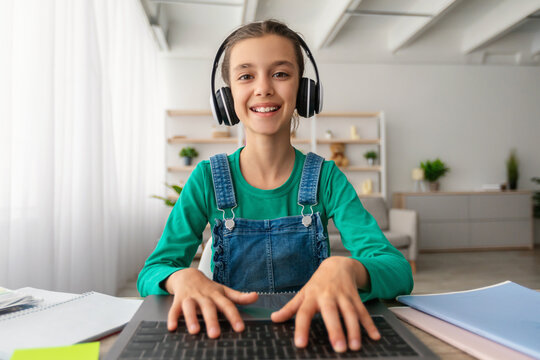 Portrait of cute little girl in headset using laptop, studying online at home, interested happy child typing on keyboard looking at pc screen, watching webinar, online course, webcam point of view - Powered by Adobe