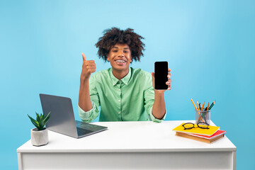 Cheerful black student guy holding smartphone with blank screen and showing thumb up, sitting at table over blue background. Teen recommending website for learning online