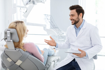 Obraz premium Male dentist showing female patient jaw x-ray on digital tablet, dental clinic interior. Bearded man stomatologist having conversation with lady in dental chair, holding pad, side view, copy space