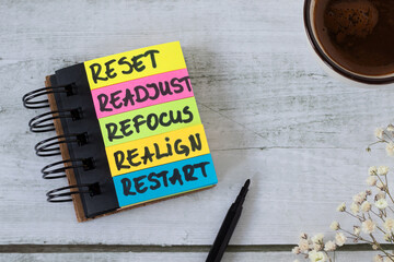 Reset, readjust, refocus, realign, restart, inspirational text in notebook with coffee cup on wooden background. Top view. New beginning, progress, goals, success, and aspirations concept.