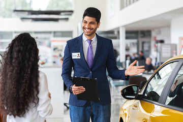 Wealthy middle-eastern young woman choosing new car, having conversation with friendly sales...