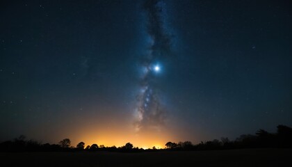 Night sky above dark horizon with luminous Milky Way. Distant town lights create orange glow under vast starry expanse. Trees silhouette against vibrant celestial view.
