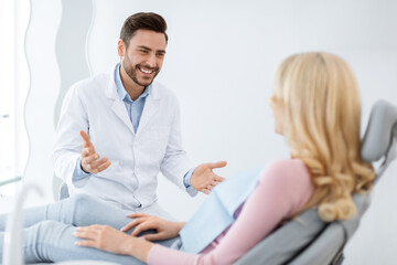Obraz premium Friendly dentist handsome millennial man in workwear having conversation with blonde woman patient sitting at dental chair, smiling and gesturing, side view. Doctor and patient relationship