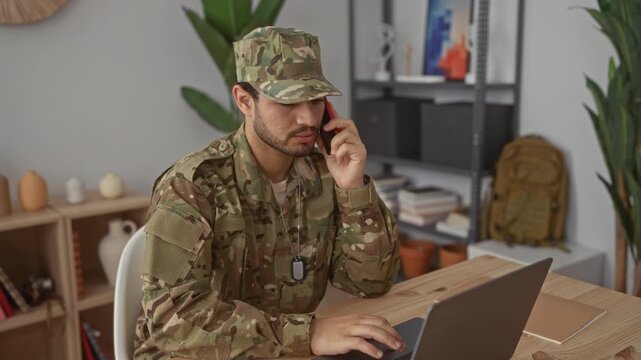 Man in camouflage uniform speaks on smartphone and types on laptop at a wooden desk in house; duty discipline.