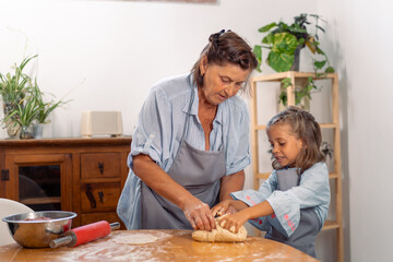 Grandmother guiding granddaughter to roll dough on kitchen table while baking together at home and enjoying fun family cooking activity