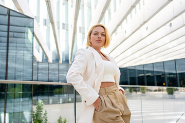 Young businesswoman with short blond hair in blazer and pants leaning on railing while looking at camera outside modern office building during daytime