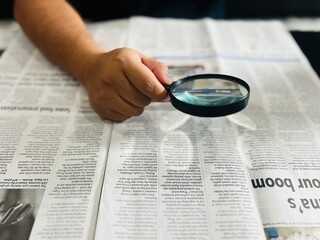 A person's hand moves a magnifying glass over a printed newspaper to read fine print, symbolizing investigative journalism, research, information seeking, or vision assistance.