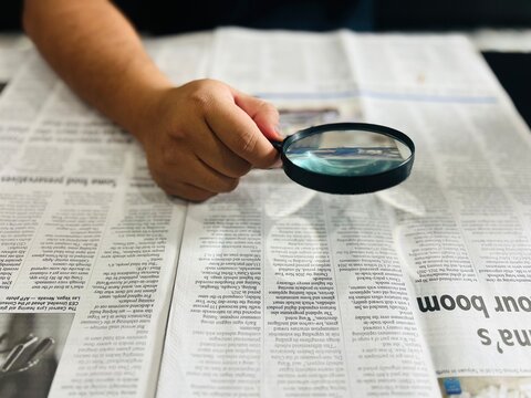 A person's hand moves a magnifying glass over a printed newspaper to read fine print, symbolizing investigative journalism, research, information seeking, or vision assistance.