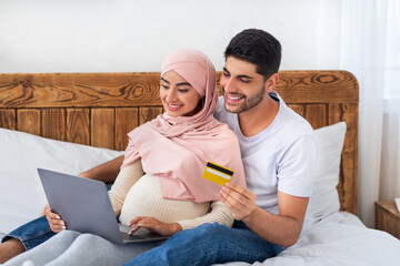 Happy arab pregnant couple shopping online at home, using laptop computer and credit card, getting ready for parenthood, sitting on bed and smiling, empty space