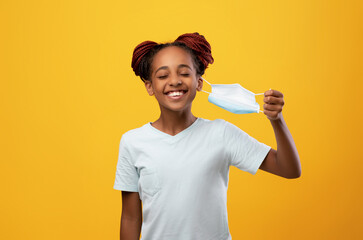 Smiling black girl teenager with closed eyes removing protective face mask, yellow studio...