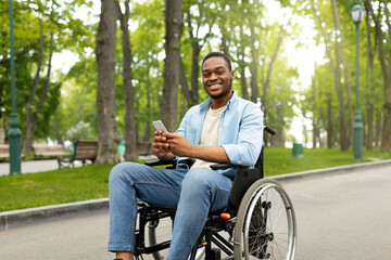 Obraz premium Happy disabled black man in wheelchair using smartphone, checking messages online at park. Young African American handicapped guy with mobile phone posting in social network, browsing web outdoors