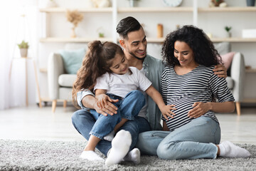 Happy middle eastern family father, pregnant mother and preschool kid posing together, sitting on floor at home, waiting for baby, cute little girl touching her expecting mom big tummy, copy space