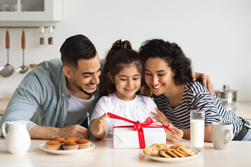 Young cheerful arabic parents giving birthday girl gift box while having breakfast at kitchen, happy adorable little kid daughter opening festive present, having celebration with mother and father