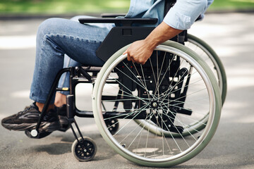 Obraz premium Unrecognizable young black man in wheelchair going for walk in city park, closeup of legs. Cropped view of millennial African American guy with physical disability spending time outdoors