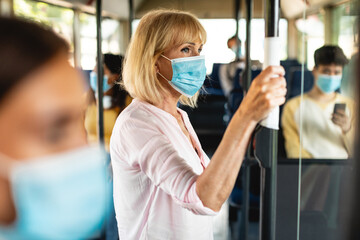Healthcare And Epidemic Concept. Elderly female passenger in protective face mask holding on handrails using tissue in urban bus, avoid touch dirty handle. City trip in context of covid pandemic