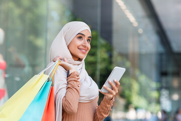 Smiling pretty millennial arabian lady in hijab enjoys online shopping with bags, surfing on phone near mall and look at empty space. New mobile app for messages from store about discount and sale