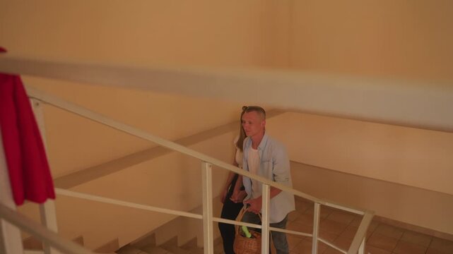 couple walking down staircase inside loft, warm beige interior with metal railing, red coat draped over banister, woman carrying woven bag, man in light shirt guiding partner, soft ambient lighting