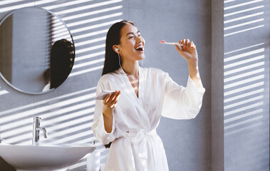 A woman stands in a bathroom with natural light shining through the window. She brushes her teeth while listening to music through her earbuds and holding a phone.