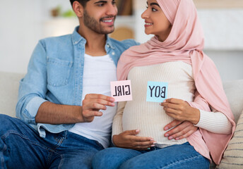 Gender reveal concept. Happy arab couple holding boy and girl sticker cards in pink and blue colors, sitting on couch at home, loving pregnant muslim spouses enjoying future parenthood