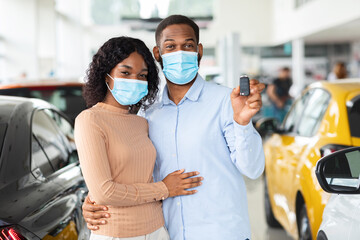 Young African American Spouses In Medical Masks Demonstrating Car Key At Camera, Millennial Black Couple Purchasing New Automobile In Dealership Office During Coronavirus Pandemic, Free Space
