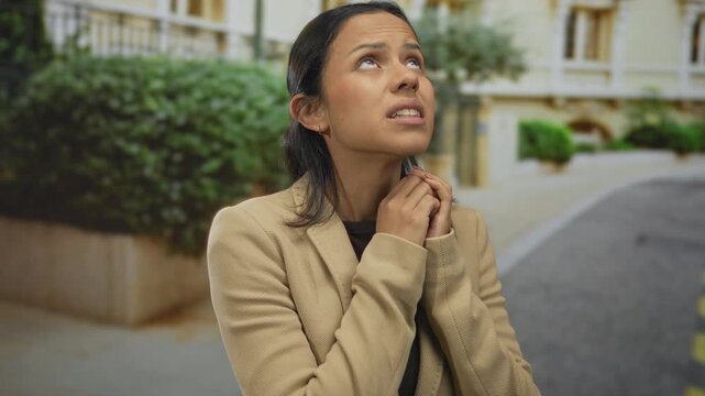 Hispanic woman in a beige jacket appears anxious standing on a quiet street, outdoors, her expression conveys fear and worry, surrounded by greenery and elegant architecture.