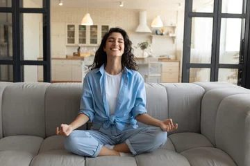 Fototapete Rund Lotusblume Cheerful young lady sitting on comfy couch, meditating with closed eyes at home, full length. Peaceful millennial woman doing yoga in lotus pose on sofa, finding inner balance, managing stress indoors  © Prostock-studio