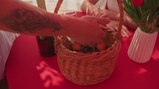 sunlit red table picnic basket champagne bottle two glasses wicker basket filled with mandarins hands reaching inside tulip vase nearby warm summer glow shadow patterns intimate couple date